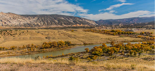 A green river  with a mountain range in the backgound and fall-colored leafy trees in the foreground 