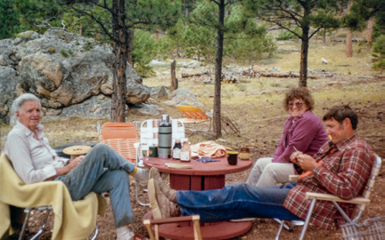 White-haired Colonel Rogers in blanket-covered lawn chair sitting with two other people in a clearin in a semi-forested area 